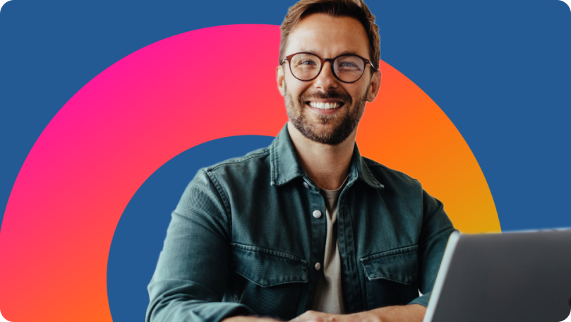 Professional white man with glasses, beard, and denim shirt looking up from laptop and smiling.